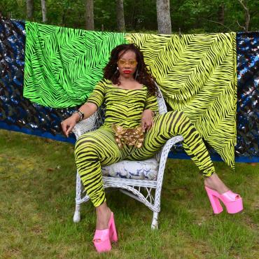Black woman in neon green zebra print body suit sitting with legs open and hot pink stacked heels on and a golden pelvis bone placed at her crotch.